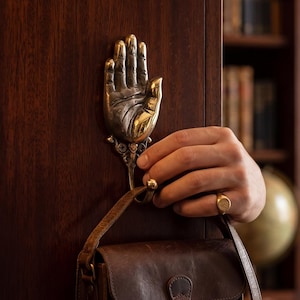 May include: A metallic wall hook shaped like an open human hand with detailed fingers and palm, mounted on a dark wood surface. The hook features an ornate base and supports a brown leather bag with a strap hanging from the wrist area of the hand.