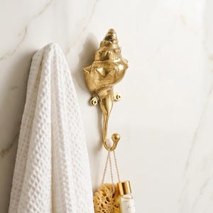 May include: A gold seashell hook fixed to a white marble wall. A white waffle-weave towel hangs to the left of the hook. A loofah and a small bottle are suspended from the hook. The Mitos Bali Studio logo is in the lower left corner.