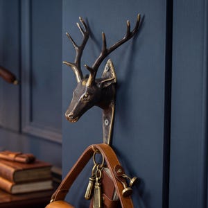 Puede incluir: Un gancho de pared con cabeza de ciervo de color bronce con una bolsa de cuero marrón colgando. El gancho está montado en una pared azul oscuro. Una lámpara de escritorio de latón y un escritorio con libros están en el fondo.