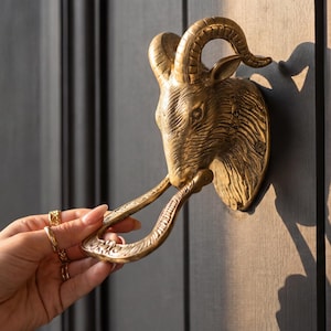 May include: A brass door knocker in the shape of a ram's head. The knocker is mounted on a dark gray wooden door. The ram's head has detailed features, including horns and a textured surface. The knocker is being held by a hand.