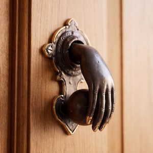 May include: A dark bronze door knocker mounted on a light brown wooden door. The knocker features a sculpted hand reaching out to grasp a spherical ball, set against an ornate, vertically elongated metal backplate with visible mounting screws.