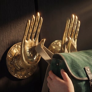 Puede incluir: Dos perchas de pared doradas en forma de mano con detalles intrincados están montadas sobre una superficie de madera oscura. Una bolsa de lona verde con correas de cuero cuelga de una percha. Las perchas añaden un elemento decorativo único.
