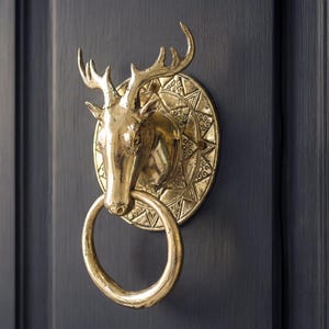 May include: A brass door knocker featuring a stag's head design, with a circular backplate adorned with geometric patterns. The knocker has a ring handle and is mounted on a dark blue door.