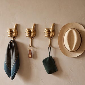 Pode incluir: Três ganchos de parede de metal dourado em forma de mãos fazendo um gesto de polegar para cima. Cada gancho possui um braço de metal voltado para baixo para pendurar itens. Um lenço estampado, um porta-chaves de couro e uma bolsa pequena.