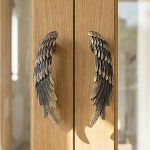 May include: Wooden cabinet doors featuring bronze angel wing handles. The handles are intricately detailed with feather patterns. The doors are set against a bright, blurred background, with a glimpse of a window and curtains.