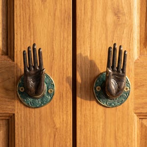 May include: Two bronze-toned hand-shaped cabinet pulls mounted on a wooden cabinet. Each hand is attached to a circular, teal-colored base with a decorative pattern. The cabinet doors are made of light-colored wood.