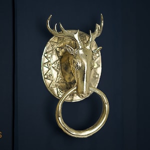Op de afbeelding: Een gouden deurklopper in de vorm van een hertenkop met geweien. De klopper heeft een ronde basis met een ingewikkeld ontwerp en een ring om aan te trekken.