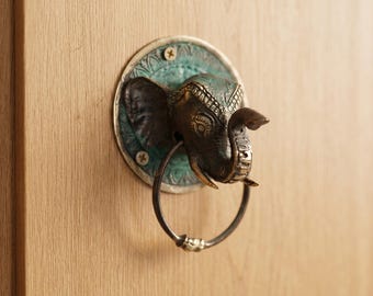 May include: An antique-style door knocker featuring an elephant head design. The knocker is made of dark metal with intricate detailing, mounted on a round, teal-colored plate. The door knocker has a ring for knocking and is attached to a wooden door.