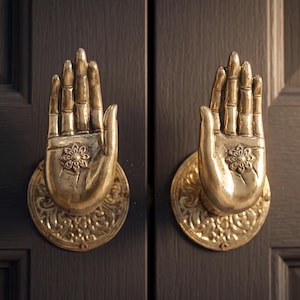 May include: Two brass-colored door knockers shaped like open hands, mounted on a dark brown door. Each hand features intricate detailing and a floral design on the palm, set on a decorative circular base. The door has a paneled design.