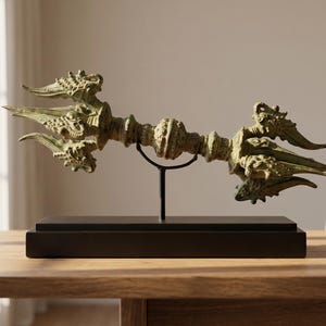 May include: A bronze-coloured vajra, a ritual object, is displayed on a black stand atop a wooden table. The vajra features intricate dragon-like carvings and a central orb, showcasing detailed craftsmanship.
