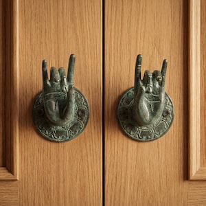 Peut inclure: Deux heurtoirs de porte en bronze en forme de mains faisant le signe 'rock on', montés sur une porte en bois clair. Les heurtoirs présentent un aspect vieilli et détaillé, ajoutant un élément décoratif unique.