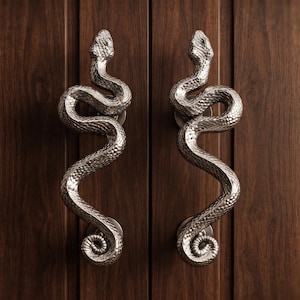 Puede incluir: Dos manijas de puerta de metal plateado con forma de serpientes enroscadas con escamas texturizadas. Las manijas están montadas verticalmente en una puerta de madera marrón oscuro con veta visible, presentando un cuerpo curvo y una cola en espiral.
