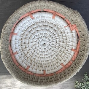 May include: A round wall hanging made of woven rope with a beige outer ring and a white centre. The outer ring has a peach-coloured band around it.