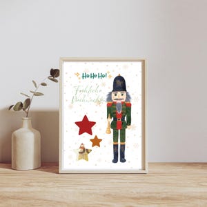 May include: Framed print featuring a watercolor illustration of a nutcracker soldier in a green and red uniform, holding a trumpet. The print includes the text "Ho Ho Ho!" and "Fröhliche Weihnachten" with red and gold stars.