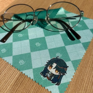 May include: Round, silver-framed eyeglasses with brown temples rest on a green and white checkered cleaning cloth. The cloth features a cartoon character illustration. The glasses have clear lenses.
