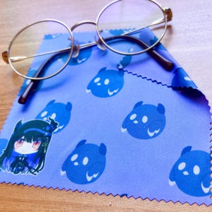 May include: A blue microfiber cleaning cloth with a repeating pattern of blue cartoon ghosts and a single cartoon character with black hair and a black and white outfit. A pair of round silver-rimmed glasses are lying on top of the cloth.