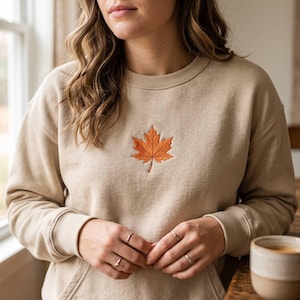 Könnte beinhalten: Ein beigefarbenes Sweatshirt mit Rundhalsausschnitt und einer mittig platzierten, gestickten orangefarbenen Ahornblatt-Applikation. Das Kleidungsstück hat lange Ärmel mit gerippten Bündchen und einen gerippten Halsausschnitt für einen schlichten Look.