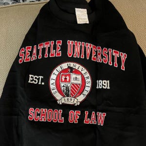 Puede incluir: Sudadera negra con letras rojas y blancas que dicen "SEATTLE UNIVERSITY" y "SCHOOL OF LAW". Un emblema circular en el centro muestra el nombre de la universidad y "1891". La sudadera es de estilo cuello redondo cl&aacute;sico.