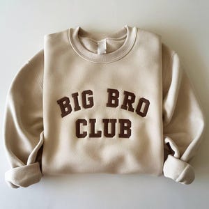 Puede incluir: Sudadera beige con las palabras "BIG BRO CLUB" en letras marrones. La sudadera de cuello redondo tiene mangas largas con pu&ntilde;os enrollados. La tela parece suave y c&oacute;moda.