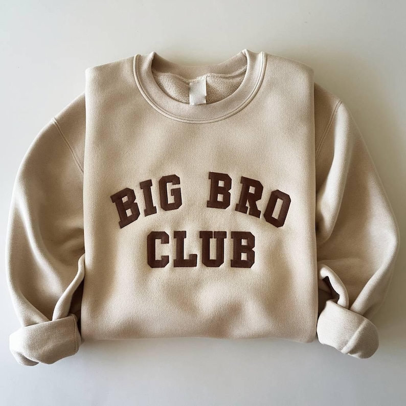 Puede incluir: Sudadera beige con las palabras "BIG BRO CLUB" en letras marrones. La sudadera de cuello redondo tiene mangas largas con pu&ntilde;os enrollados. La tela parece suave y c&oacute;moda.