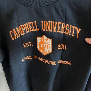 Puede incluir: Sudadera negra con texto bordado en naranja "CAMPBELL UNIVERSITY", "EST. 2011" y "SCHOOL OF OSTEOPATHIC MEDICINE". El dise&ntilde;o incluye un escudo en forma de escudo en naranja y blanco.