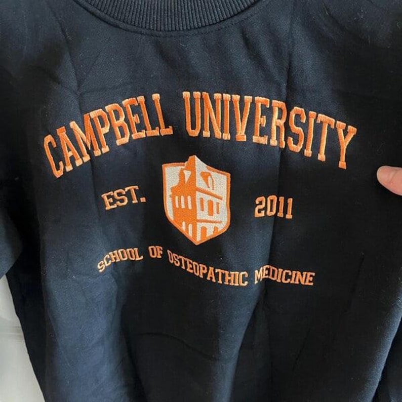 Puede incluir: Sudadera negra con texto bordado en naranja "CAMPBELL UNIVERSITY", "EST. 2011" y "SCHOOL OF OSTEOPATHIC MEDICINE". El dise&ntilde;o incluye un escudo en forma de escudo en naranja y blanco.