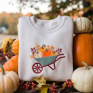 Könnte beinhalten: Weißes Sweatshirt mit einem gestickten Design einer tealfarbenen Schubkarre, gefüllt mit Kürbissen und Herbstblättern. Die Schubkarre hat ein rotes Rad und einen Griff. Das Design ist auf der Vorderseite zentriert und erinnert an die Herbsternte.