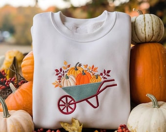 May include: White sweatshirt with an embroidered design of a teal wheelbarrow filled with pumpkins and autumn leaves. The wheelbarrow has a red wheel and handle. The design is centered on the front, evoking a fall harvest theme.