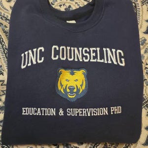 Puede incluir: Sudadera azul marino con el texto blanco "UNC COUNSELING". Debajo, un logotipo de oso amarillo con contorno azul y el texto "EDUCATION & SUPERVISION PHD". La sudadera est&aacute; doblada.