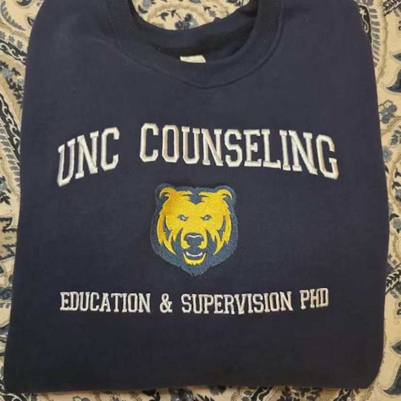 Puede incluir: Sudadera azul marino con el texto blanco "UNC COUNSELING". Debajo, un logotipo de oso amarillo con contorno azul y el texto "EDUCATION & SUPERVISION PHD". La sudadera est&aacute; doblada.