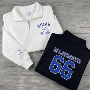Può includere: Due felpe con zip a un quarto di collo. Una è bianca con "Briar University" e bastoni da hockey incrociati ricamati in blu. L'altra è nera con "Di Laurentis" e il numero "66" in blu. Entrambe hanno un colletto alto.