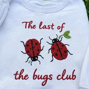 May include: White sweatshirt with embroidered red ladybugs and the text "The last of the bugs club" in red script. The ladybugs have black spots and antennae. A green leaf is also embroidered.