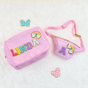 Puede incluir: Dos bolsas de tela rosa con letras de colores y parches de arcoíris. La bolsa más grande dice "LINDA" y la bolsa más pequeña dice "A". Ambas bolsas tienen una textura suave y esponjosa.