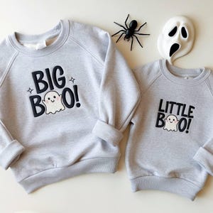 May include: Two light gray sweatshirts with the text "BIG BOO!" and "LITTLE BOO!" in black, each featuring a ghost graphic. A black spider and a white ghost mask are also present, suggesting a Halloween theme.