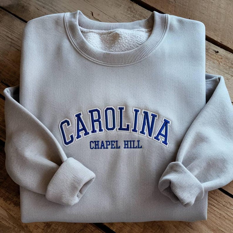 Op de afbeelding: Een lichtgrijs sweatshirt met het woord "CAROLINA" in blauwe letters over de borst, met daaronder "CHAPEL HILL". De sweatshirt heeft een ronde hals en manchetten, gepresenteerd op een houten oppervlak.