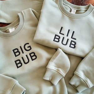 May include: Two sage green sweatshirts with black embroidered text. One sweatshirt says "BIG BUB" and the other says "LIL BUB".