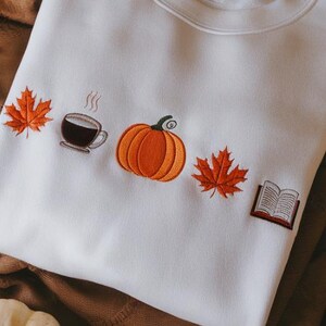 May include: Cream-coloured sweatshirt with embroidered autumnal motifs. Includes an orange pumpkin, a coffee cup, two orange maple leaves, and an open book. Ideal for autumn.