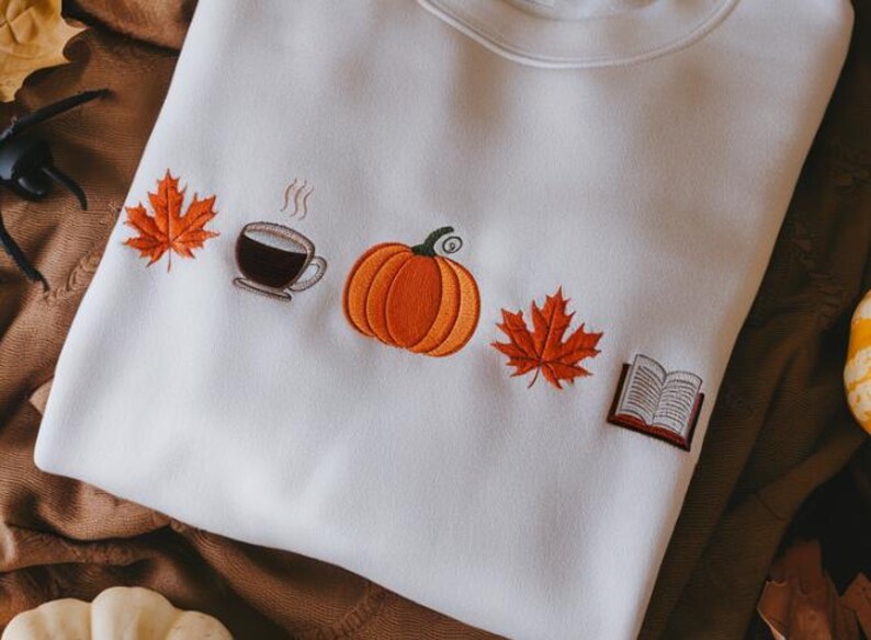 May include: Cream-coloured sweatshirt with embroidered autumnal motifs. Includes an orange pumpkin, a coffee cup, two orange maple leaves, and an open book. Ideal for autumn.