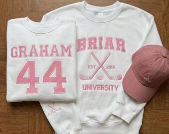 May include: White sweatshirts with pink embroidered text and graphics. One sweatshirt displays "GRAHAM 44", the other reads "GRAHAM BRIAR UNIVERSITY EST 2015" with crossed hockey sticks. A pink baseball cap with a hockey stick logo completes the set.