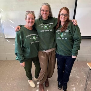Puede incluir: Tres personas con sudaderas verde oscuro con el texto "SOUTHEASTERN LOUISIANA UNIVERSITY EST. 1925 MASTER OF SOCIAL WORK" y un gran logotipo "S". Est&aacute;n en una habitaci&oacute;n con una pizarra y una mesa.