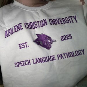 Puede incluir: Sudadera blanca con texto bordado en morado: "Abilene Christian University", "Speech Language Pathology", "Est. 2025" y un logotipo de pantera morada. La sudadera es de cuello redondo.