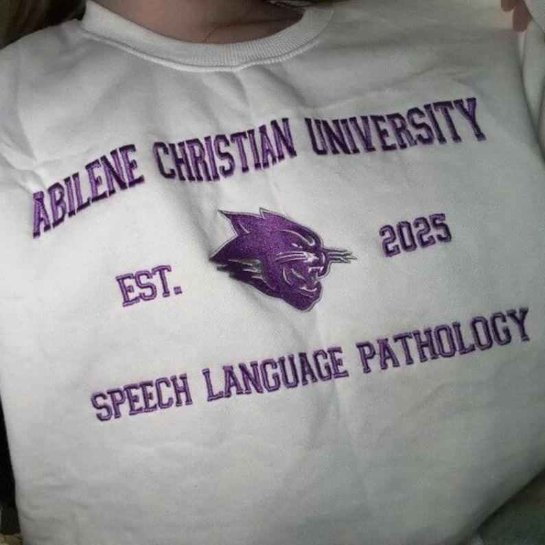 Puede incluir: Sudadera blanca con texto bordado en morado: "Abilene Christian University", "Speech Language Pathology", "Est. 2025" y un logotipo de pantera morada. La sudadera es de cuello redondo.
