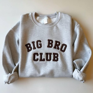 Puede incluir: Una sudadera gris claro con las palabras "BIG BRO CLUB" en letras marr&oacute;n oscuro, estilo universitario. La sudadera tiene cuello redondo y mangas largas con pu&ntilde;os enrollados. La prenda est&aacute; hecha de un material suave.