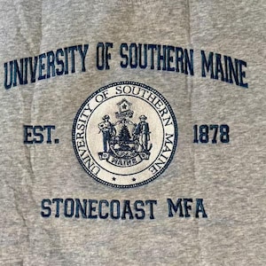 Puede incluir: Una sudadera gris con las palabras "UNIVERSITY OF SOUTHERN MAINE" en azul marino arqueadas en la parte superior. Debajo hay un emblema circular con las palabras "UNIVERSITY OF SOUTHERN MAINE" y el a&ntilde;o "1878". El texto "STONECOAST MFA" est&aacute; en la parte inferior.