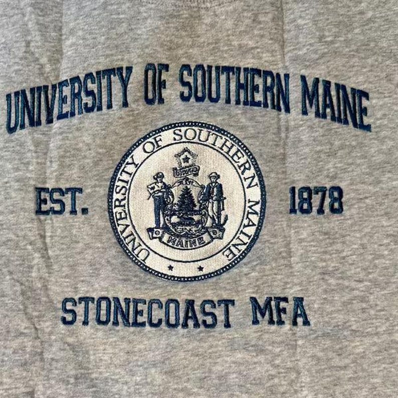 Puede incluir: Una sudadera gris con las palabras "UNIVERSITY OF SOUTHERN MAINE" en azul marino arqueadas en la parte superior. Debajo hay un emblema circular con las palabras "UNIVERSITY OF SOUTHERN MAINE" y el a&ntilde;o "1878". El texto "STONECOAST MFA" est&aacute; en la parte inferior.