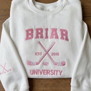 May include: White sweatshirt with pink embroidered text and graphics. The word "BRIAR" is arched across the top, with crossed hockey sticks and a puck below. The text "EST 2015" is on the left and right of the crossed sticks, and "UNIVERSITY" is below.