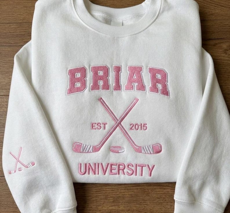 May include: White sweatshirt with pink embroidered text and graphics. The word "BRIAR" is arched across the top, with crossed hockey sticks and a puck below. The text "EST 2015" is on the left and right of the crossed sticks, and "UNIVERSITY" is below.