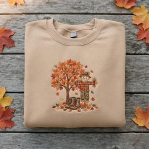 May include: A beige crewneck sweatshirt featuring an embroidered design of a scarecrow standing next to a large tree with orange and red autumn leaves. The scarecrow wears a plaid shirt and brown boots, surrounded by fallen leaves on the ground.