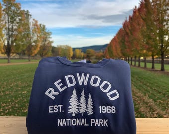 May include: A navy blue sweatshirt with the word "REDWOOD" embroidered in white, along with "EST. 1968" and "NATIONAL PARK". The sweatshirt is folded on a wooden surface, with a background of trees and a blue sky.