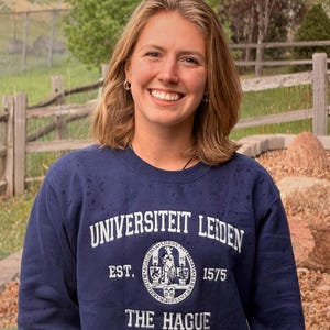 Puede incluir: Una mujer con una sudadera azul marino con las palabras "UNIVERSITEIT LEIDEN" y "THE HAGUE" impresas en blanco. La sudadera tiene un emblema circular con texto y una figura. La mujer sonr&iacute;e, con un fondo natural al aire libre.